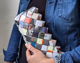 May include: A woven clutch made from recycled paper, featuring a geometric pattern of colorful squares. The clutch is held against a denim jacket, showcasing its unique texture and design. The color palette includes shades of orange, gray, and white.