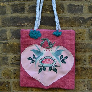 May include: Pink tote bag with a heart-shaped design featuring two blue birds and a dish. The bag has white and blue handles and teal pom-poms. The bag is made of a woven material.