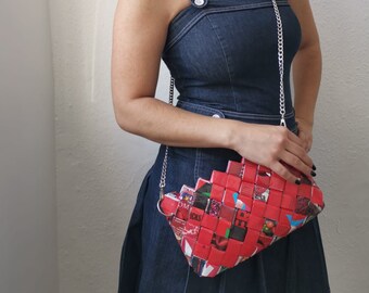May include: A red woven clutch with a silver chain strap. The clutch is made from recycled materials and features a geometric pattern. The person in the image is wearing a denim dress.