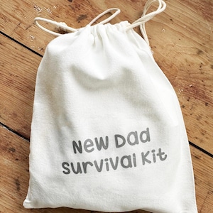 May include: A white drawstring bag with the text "New Dad Survival Kit" printed in grey. The bag is made of a textured fabric and sits on a wooden surface. The drawstring is a light beige colour.