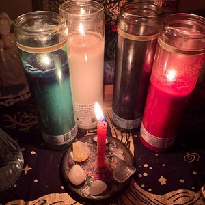 May include: A collection of lit candles in glass jars, including green, white, black, and red. A small red candle burns in a glass dish with crystals and stones. The scene is set on a dark, patterned surface.
