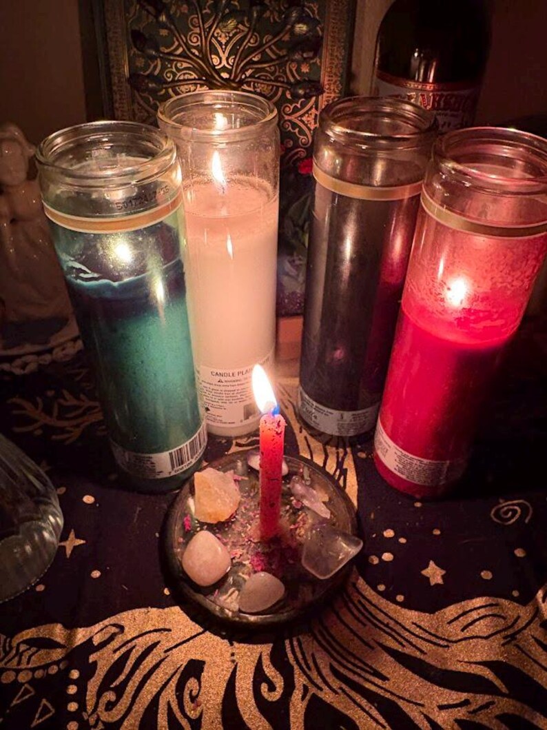 May include: A collection of lit candles in glass jars, including green, white, black, and red. A small red candle burns in a glass dish with crystals and stones. The scene is set on a dark, patterned surface.