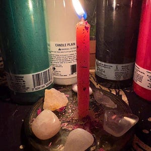 May include: A lit red candle in a metal dish with crystals and dried flower petals. Several large, cylindrical candles in green, white, black, and red are in the background. The white candle has the text "CANDLE PLAIN".