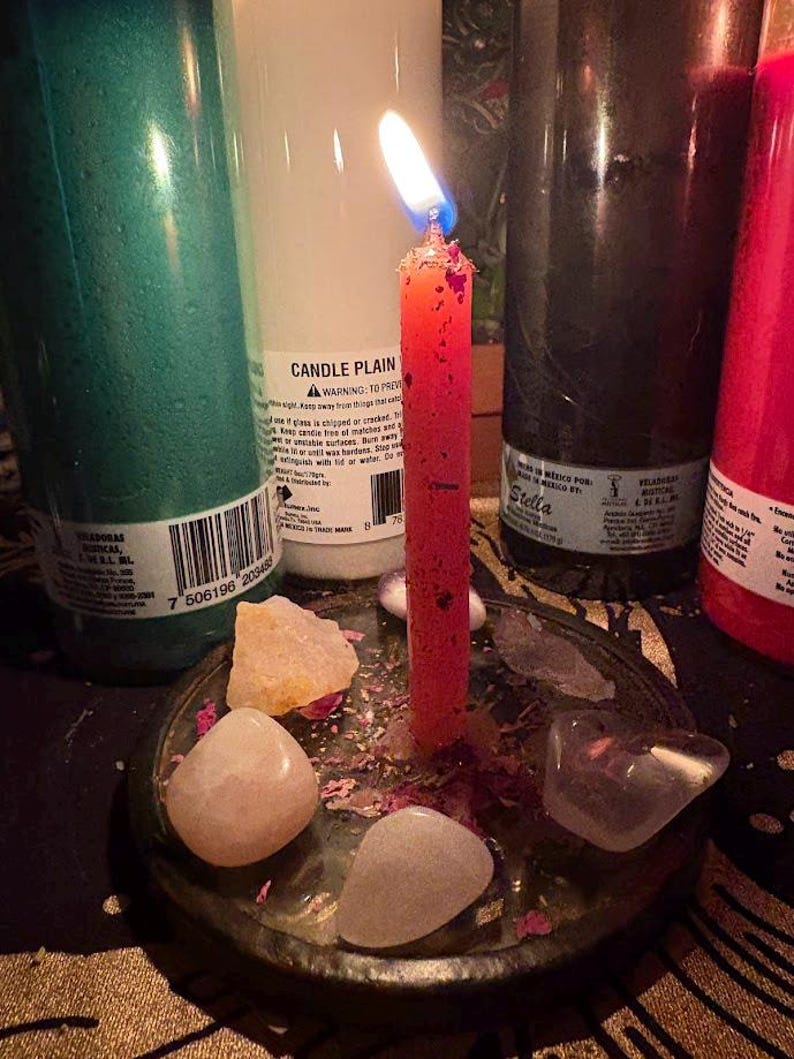May include: A lit red candle in a metal dish with crystals and dried flower petals. Several large, cylindrical candles in green, white, black, and red are in the background. The white candle has the text "CANDLE PLAIN".