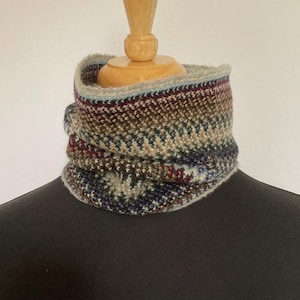 May include: A multi-colored knitted cowl in shades of blue, brown, and burgundy. The cowl is displayed on a mannequin and features a textured pattern. The neck warmer is designed to be worn around the neck for warmth and style.