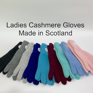 May include: A collection of cashmere gloves arranged in a row, featuring colours including black, grey, navy, teal, burgundy, light blue, and pink. Each pair has a small circular white tag on the cuff. Text reads: Ladies Cashmere Gloves Made in Scotland.