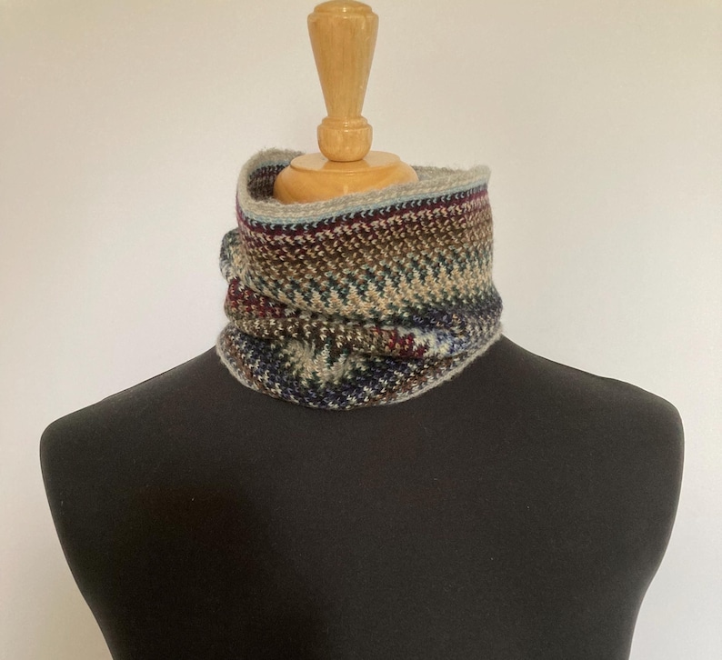 May include: A multi-colored knitted cowl in shades of blue, brown, and burgundy. The cowl is displayed on a mannequin and features a textured pattern. The neck warmer is designed to be worn around the neck for warmth and style.