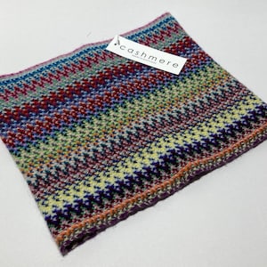 May include: A colorful cashmere neck warmer with a geometric pattern in shades of blue, red, green, and purple. The tag reads "cashmere". The accessory is folded and ready to wear.