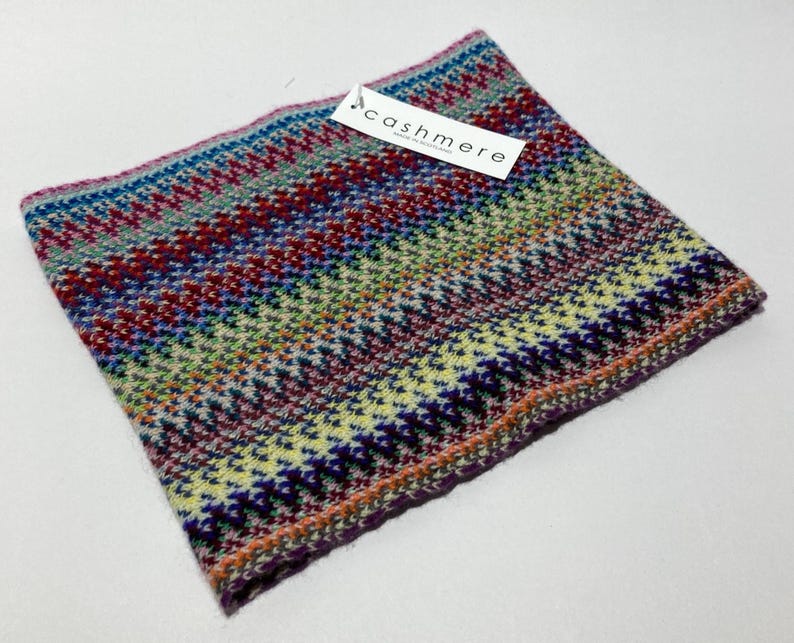 May include: A colorful cashmere neck warmer with a geometric pattern in shades of blue, red, green, and purple. The tag reads "cashmere". The accessory is folded and ready to wear.
