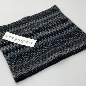 May include: A knitted cashmere neck warmer in shades of black, gray, and blue. The accessory features a geometric pattern and a tag that reads "cashmere MADE IN SCOTLAND".