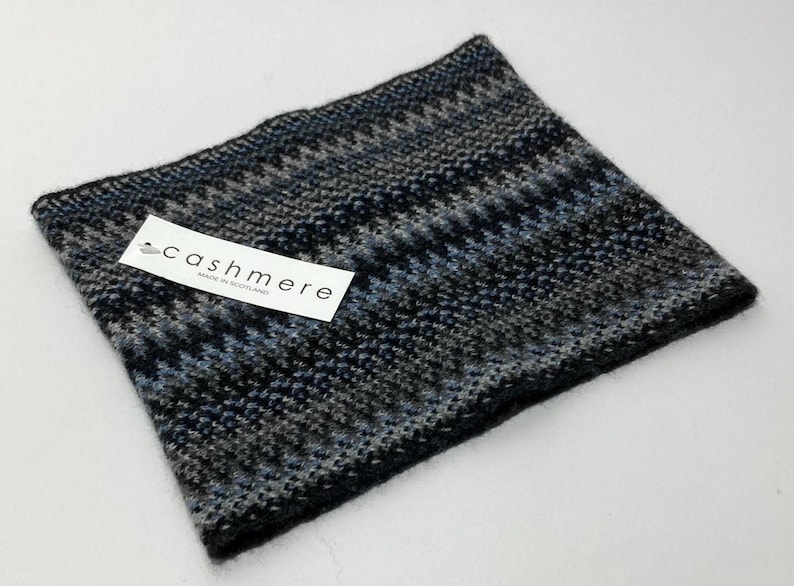 May include: A knitted cashmere neck warmer in shades of black, gray, and blue. The accessory features a geometric pattern and a tag that reads "cashmere MADE IN SCOTLAND".