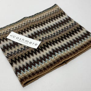 May include: A folded cashmere scarf with a geometric pattern in shades of brown, beige, and teal. The scarf has a tag that reads "cashmere made in Scotland". The scarf is a cozy accessory.