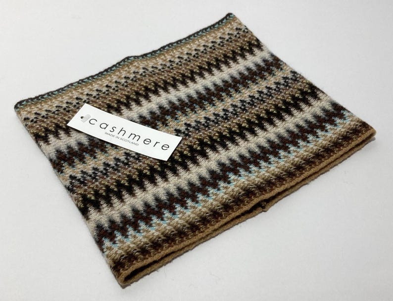 May include: A folded cashmere scarf with a geometric pattern in shades of brown, beige, and teal. The scarf has a tag that reads "cashmere made in Scotland". The scarf is a cozy accessory.