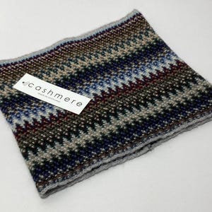 May include: A multi-colored cashmere neck warmer with a geometric pattern in shades of blue, brown, gray, and burgundy. The label reads "cashmere MADE IN SCOTLAND". The neck warmer is folded and has a gray border.