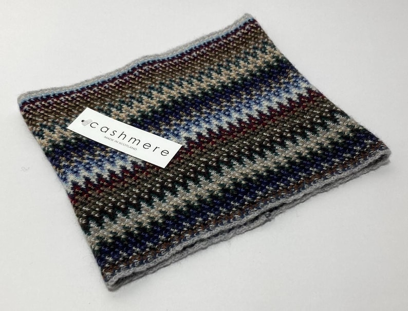 May include: A multi-colored cashmere neck warmer with a geometric pattern in shades of blue, brown, gray, and burgundy. The label reads "cashmere MADE IN SCOTLAND". The neck warmer is folded and has a gray border.