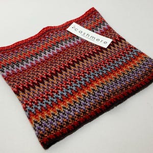 May include: A colorful cashmere cowl in shades of red, orange, purple, and black. The cowl features a geometric pattern and a tag that reads "cashmere". The knitted accessory is designed to be worn around the neck.