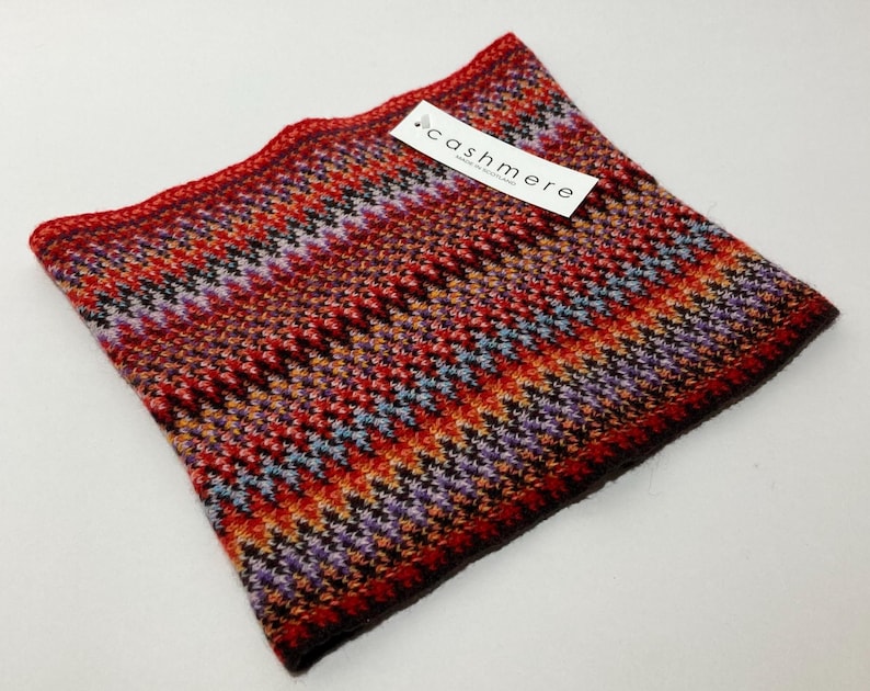 May include: A colorful cashmere cowl in shades of red, orange, purple, and black. The cowl features a geometric pattern and a tag that reads "cashmere". The knitted accessory is designed to be worn around the neck.