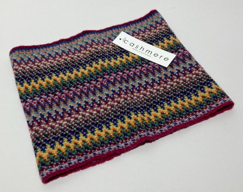 May include: A colorful cashmere neck warmer with a zig-zag pattern in shades of purple, blue, yellow, and green. The edge is a deep red. A tag with the word "cashmere" is attached.