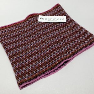 May include: A square, patterned scarf with a houndstooth design in shades of brown, red, and white. The scarf has a pink border and a tag that reads "cashmere". The scarf is made of a soft, knitted material.