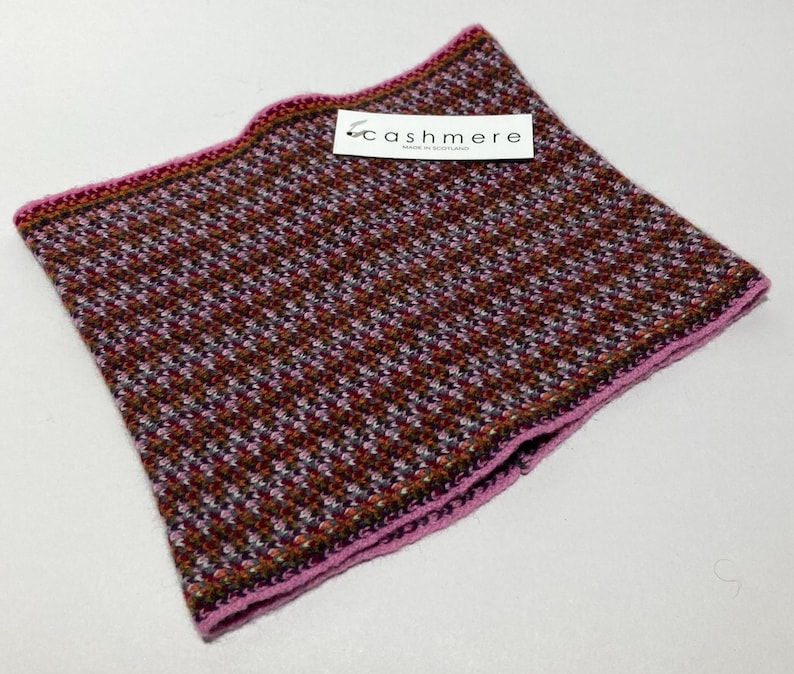 May include: A square, patterned scarf with a houndstooth design in shades of brown, red, and white. The scarf has a pink border and a tag that reads "cashmere". The scarf is made of a soft, knitted material.