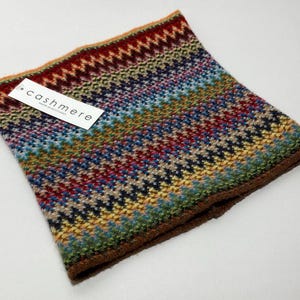 May include: A colorful cashmere neck warmer with a geometric pattern in shades of red, blue, green, and yellow. The label reads "cashmere". The edges are trimmed with a brown border.