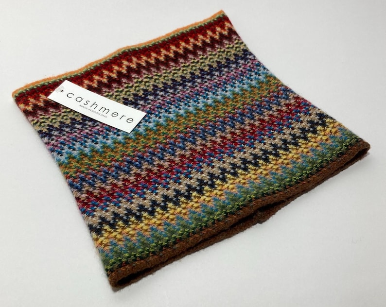 May include: A colorful cashmere neck warmer with a geometric pattern in shades of red, blue, green, and yellow. The label reads "cashmere". The edges are trimmed with a brown border.
