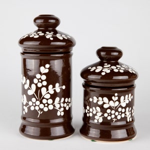 May include: Two brown ceramic canisters with white floral designs. The canisters have lids and are of different sizes.