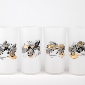 May include: Set of four frosted glass tumblers with gold trim. Each tumbler features a black and white illustration of a vintage car with the car's make and year printed below. The tumblers feature the Oldsmobile 1904, the Ford 1903, the Rambler 1903, and the Ford 1903.