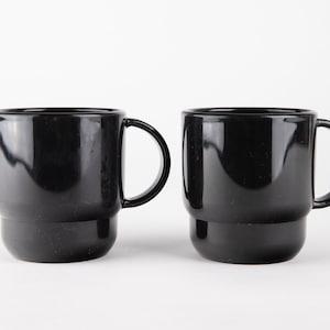 May include: Two black ceramic mugs with a simple design. The mugs have a rounded shape and a handle on the right side.