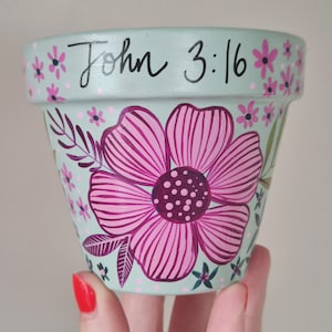 May include: A hand-painted, pale turquoise flower pot featuring a large pink flower design. The pot is decorated with smaller pink flowers and the text "John 3:16" in black script. The pot is held by a person.