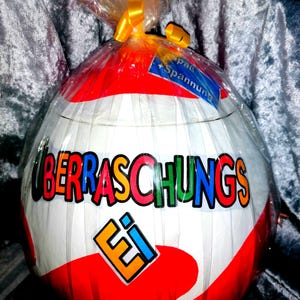 Könnte beinhalten: Ein großes, eiförmiges Objekt, verpackt in durchsichtiger Folie mit einer gelben Schleife. Die Oberfläche ist weiß mit roten Bereichen und zeigt den Schriftzug "ÜBERRASCHUNGS Ei" in bunten Buchstaben sowie ein blaues Etikett mit "Spaß + Spannung".