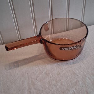 Può includere: Una casseruola Vision ambrata trasparente con un lungo manico. La casseruola ha una forma a ciotola arrotondata e un manico che si estende dal lato. Il logo Vision è visibile sul lato della pentola. Questa pentola è adatta per cucinare.