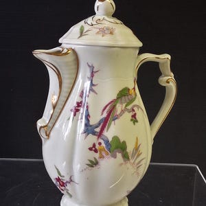 Peut inclure: Une cafetière en porcelaine blanche avec couvercle, ornée d'un motif peint à la main représentant un oiseau coloré perché sur une branche avec des feuilles vertes et des fleurs rouges. Le bord du bec, de l'anse, du couvercle et la base sont dorés.