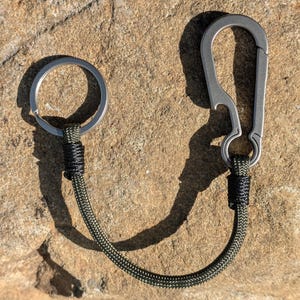 May include: A keyring featuring a silver-toned metal carabiner with an integrated bottle opener, connected by a dark olive green paracord lanyard to a circular silver-toned metal split ring. The cord is secured at both ends with black thread.