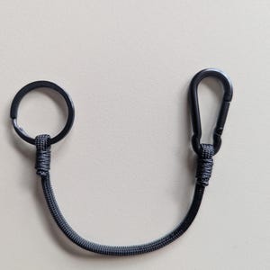 May include: A black keychain lanyard featuring a circular metal ring on one end and a spring-loaded carabiner clip on the other. The two ends are connected by a durable, woven black cord that is securely fastened to the metal hardware with tight knots.