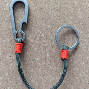 May include: A grey metal carabiner with a bottle opener feature connected by a dark green braided cord to a grey metal split key ring. The cord is secured at both ends with bright orange thread wraps, creating a durable and functional keychain.