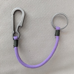 May include: A keyring featuring a grey metal carabiner with an integrated bottle opener on one end and a circular metal split ring on the other, connected by a curved length of purple paracord secured with dark green thread wrappings.