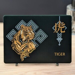 Op de afbeelding: Een rechthoekige zwarte plaat met een goudkleurige tijgerillustratie bestaande uit printplaatpatronen. Achter de tijger bevinden zich witte geometrische lijnen. Rechts staan een Chinees karakter en het woord TIGER in goud gedrukt.