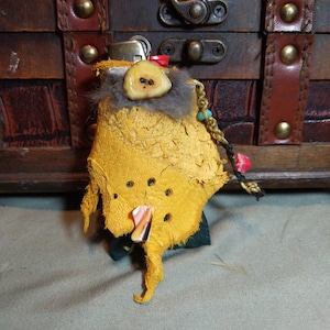 Puede incluir: Una pequeña bolsa de cuero amarilla con un amuleto de hueso y cuentas turquesas. La bolsa está decorada con una cinta roja y blanca a rayas y tiene una cremallera.