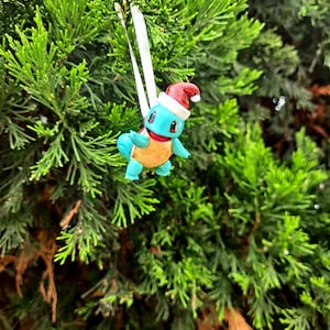 May include: A blue and yellow Squirtle Pokemon Christmas ornament wearing a red Santa hat. The ornament is hanging from a white ribbon on a green Christmas tree.