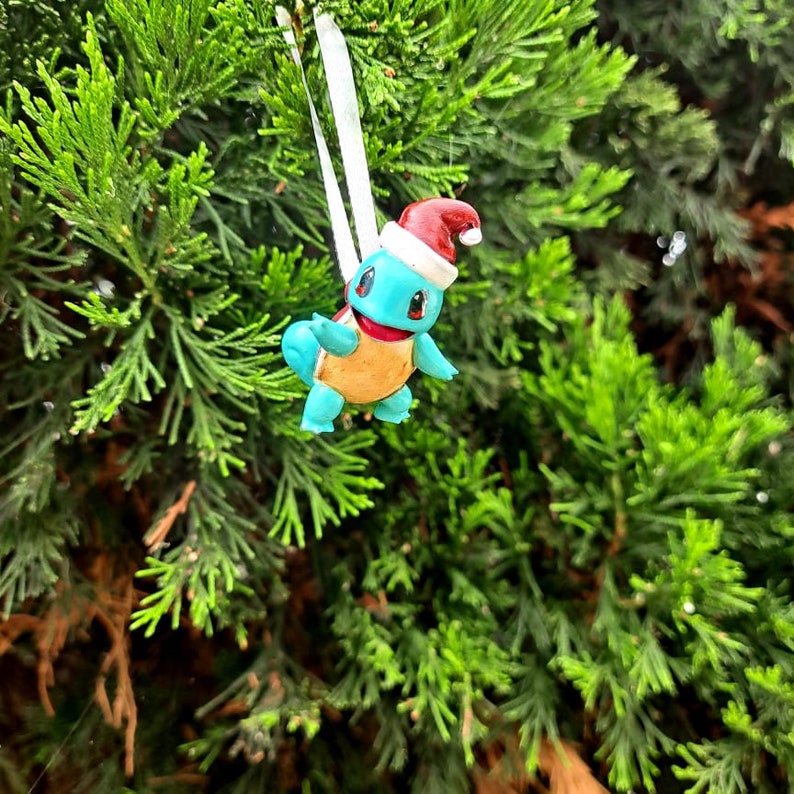 May include: A blue and yellow Squirtle Pokemon Christmas ornament wearing a red Santa hat. The ornament is hanging from a white ribbon on a green Christmas tree.