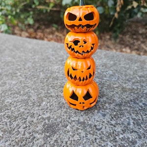 Puede incluir: Una pila de cuatro calabazas naranjas con caras talladas. Las calabazas están apiladas una encima de la otra, con la calabaza más pequeña en la parte superior. Todas las calabazas están talladas con diferentes expresiones faciales.