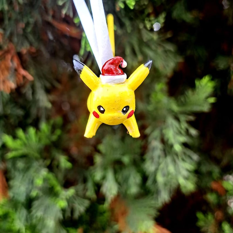 Op de afbeelding: Een gele Pikachu kerstversiering met een rode kerstmanmuts. De versiering hangt aan een wit touwtje.