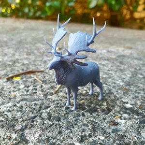 May include: A grey 3D printed figurine of a deer with large antlers. The deer is standing on a grey, textured surface.