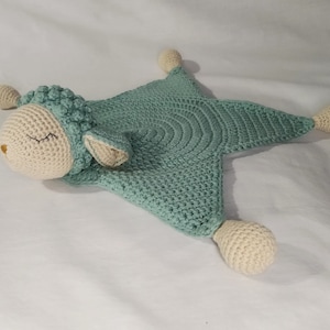 May include: A crocheted baby blanket in the shape of a sheep. The blanket is light green with a white sheep head and four white crocheted balls attached to the corners.