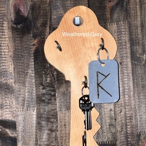 May include: Weathered grey wooden key-shaped wall hook with a metal tag with a rune symbol. The hook has two metal rings and a black cord hanging from the bottom.