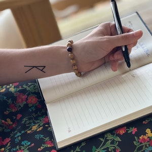 May include: A person's arm with a black tattoo of a rune on their wrist. They are wearing a wooden beaded bracelet with a dark stone bead. The person is writing in a notebook with a black pen.