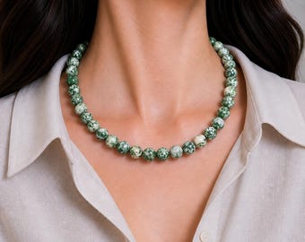 May include: A beaded necklace featuring round, speckled beads in shades of green and white. The necklace is worn around the neck, showcasing the natural stone beads. The necklace is a classic piece of jewelry.