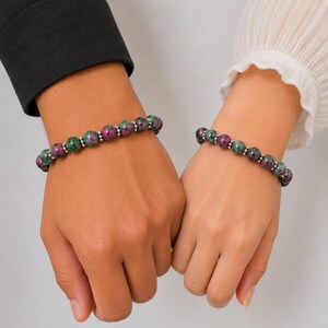 May include: Two beaded bracelets featuring round, polished ruby zoisite stones in shades of deep magenta and forest green, interspersed with small, reflective silver-toned metal spacer beads, worn on two wrists to showcase the natural stone pattern.