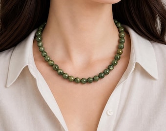 Puede incluir: Un collar de cuentas verdes redondas. El collar se lleva sobre una camisa blanca. Las cuentas tienen una superficie facetada, que refleja la luz. Una joya sencilla y elegante.